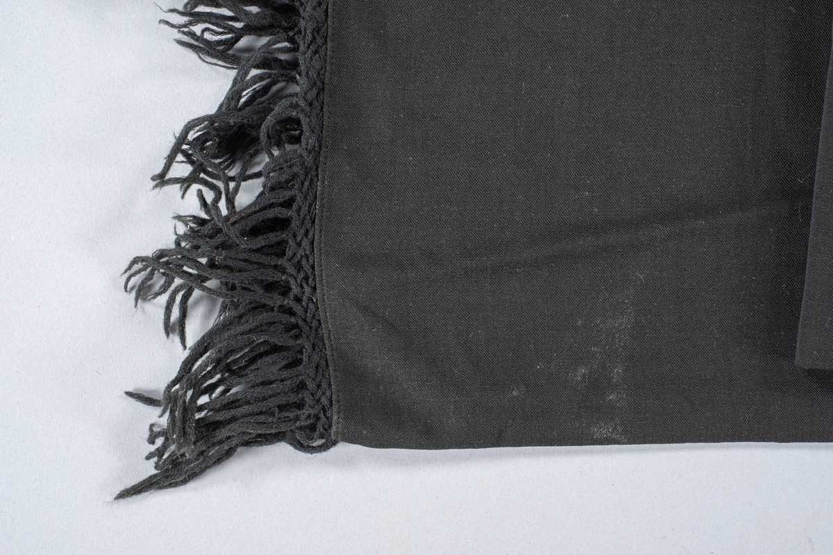 Rektangulært sort sjal i bomull. På begge kortsidene er det en flettet kant med knyttede frynser ytterst. Sjalets underside er litt lysere enn oversiden.