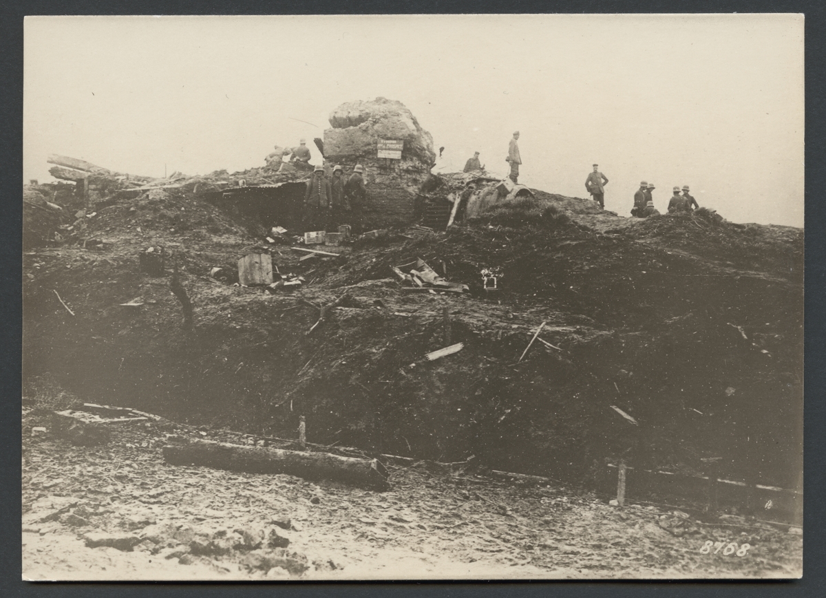 Bilden visar ett slagfält från första världskriget. Området är helt uppgrävt av artillerield och täckt med spillror av krigsmateriel. I bakgrunden syns en sönderskjuten bunker och en grupp soldater som inspekterar området.