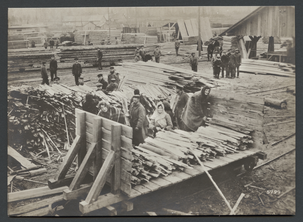 Bilden visar en lagringsplats för sågat trä. Flera arbetare håller på att lasta avfall och trärester på en godsvagn.