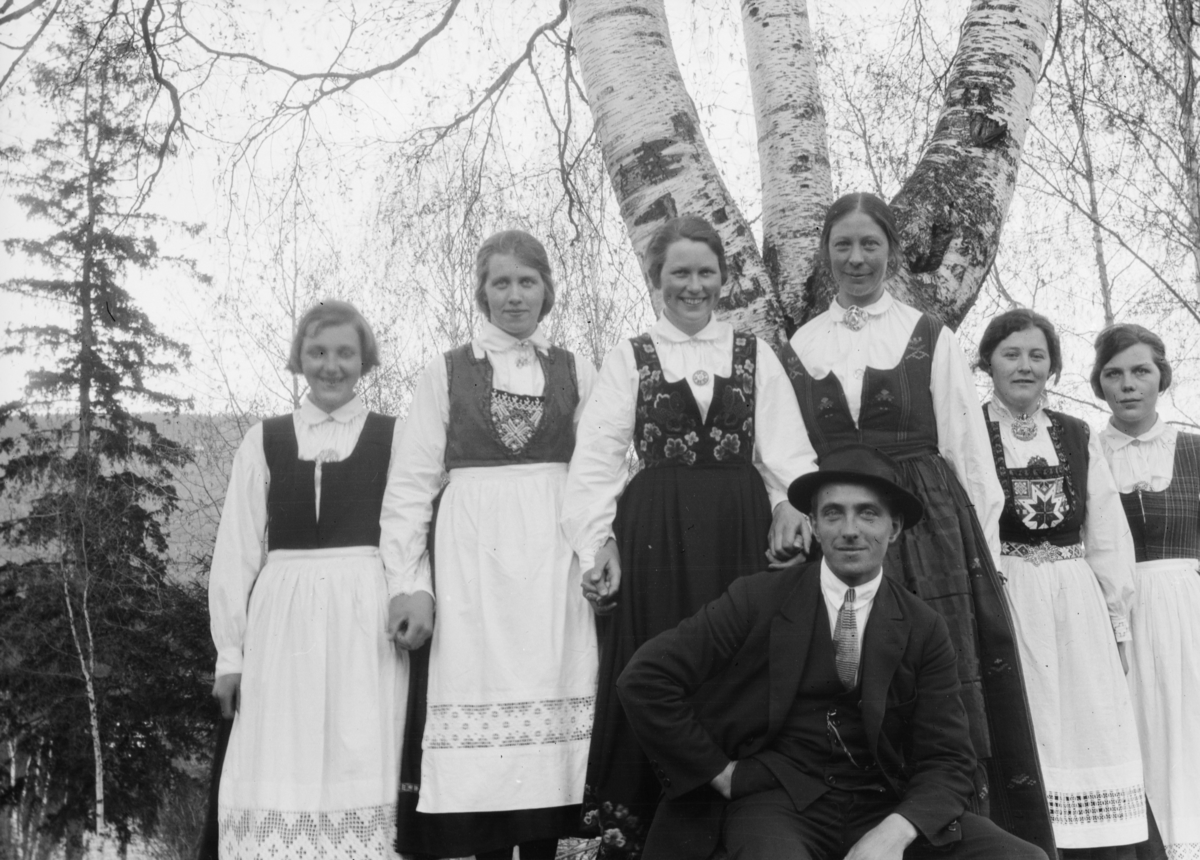 Seks unge damer med forskjellige bunader, fra blant annet Gudbrandsdalen og Hardanger. En mann. Bildet er fra bjørka ved Vonheim.