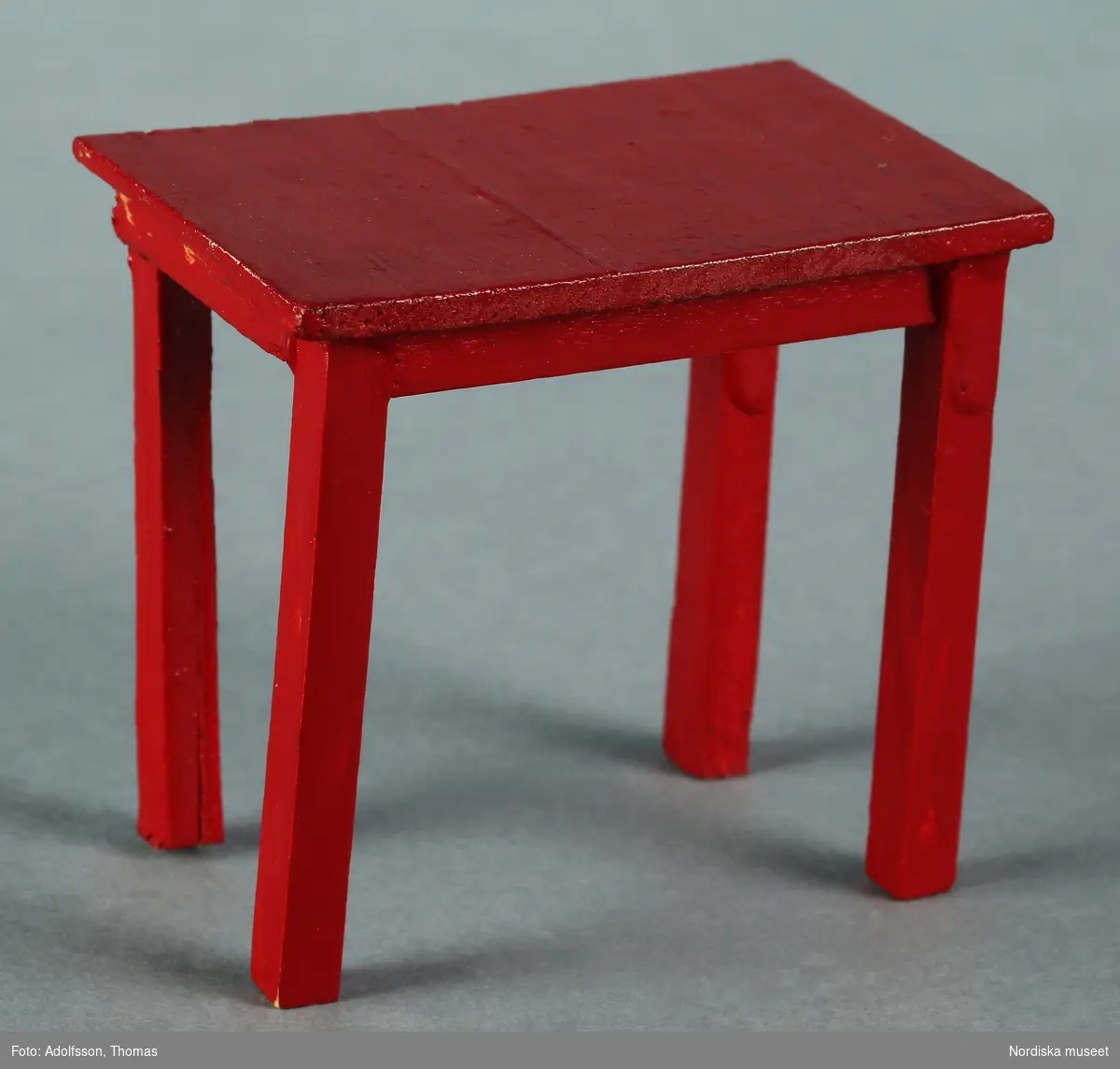 a) Ett köksbord och b-e) fyra stolar, alla av rödmålat trä, men av olika modell. b-c) Två av stolarna är raka och funktionella trästolar, troligen från 1950-talet, medan d-e) två har allmogekaraktär med utskuren dekor i ryggpartiet och blårandig klädsel. De senare är förmodligen från 1970-talet. Bordet är hemtillverkat av givarens far medan stolarna är tillverkade av Lundby. Hör till frukostrummet på första våningen i dockskåp NM.0331721+.