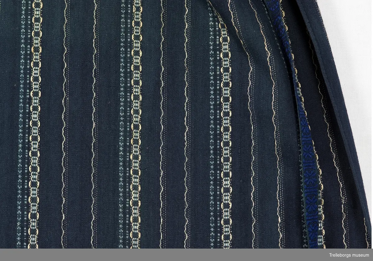 Långrandigt mörkblått midjeförkläde, hemvävt och handsytt.
Mycket tätvävt med gulbeiga och mellanblå rosengångsränder.
Kraftigt rynkat mot linningen, bredd i midjan 29 cm.
Linningen är 2 cm bred, dubbelvikt mörkblått bomullstyg med beiga ränder, vid linningens slut är mönstervävda ylleband påsydda, lysande kungsblått mot marinblå bakgrund och en tunn grön rand i kanten.168 cm långt i höger kant och 146 cm långt i vänster, 2 cm brett.
Välgjord flätad avslutning med tofsar.