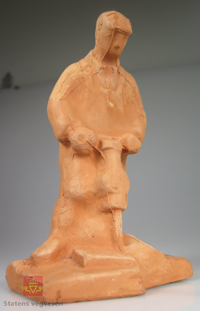 Rødbrun skulptur av en anleggsarbeider med pressluftbor. Anleggsarbeideren har et svakt omriss av klær på seg, og en lue. Detaljnivået er ellers lavt. Under er det grå filt.

Merket: N. AA. 65