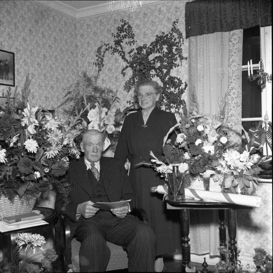 Gruppbild hemma hos Oskar Bergfoth och hans hustru Hulda Karolina. Oskar fyller 70 år och sitter på en stol omgiven av blommor. Hans hustru står bakom honom.