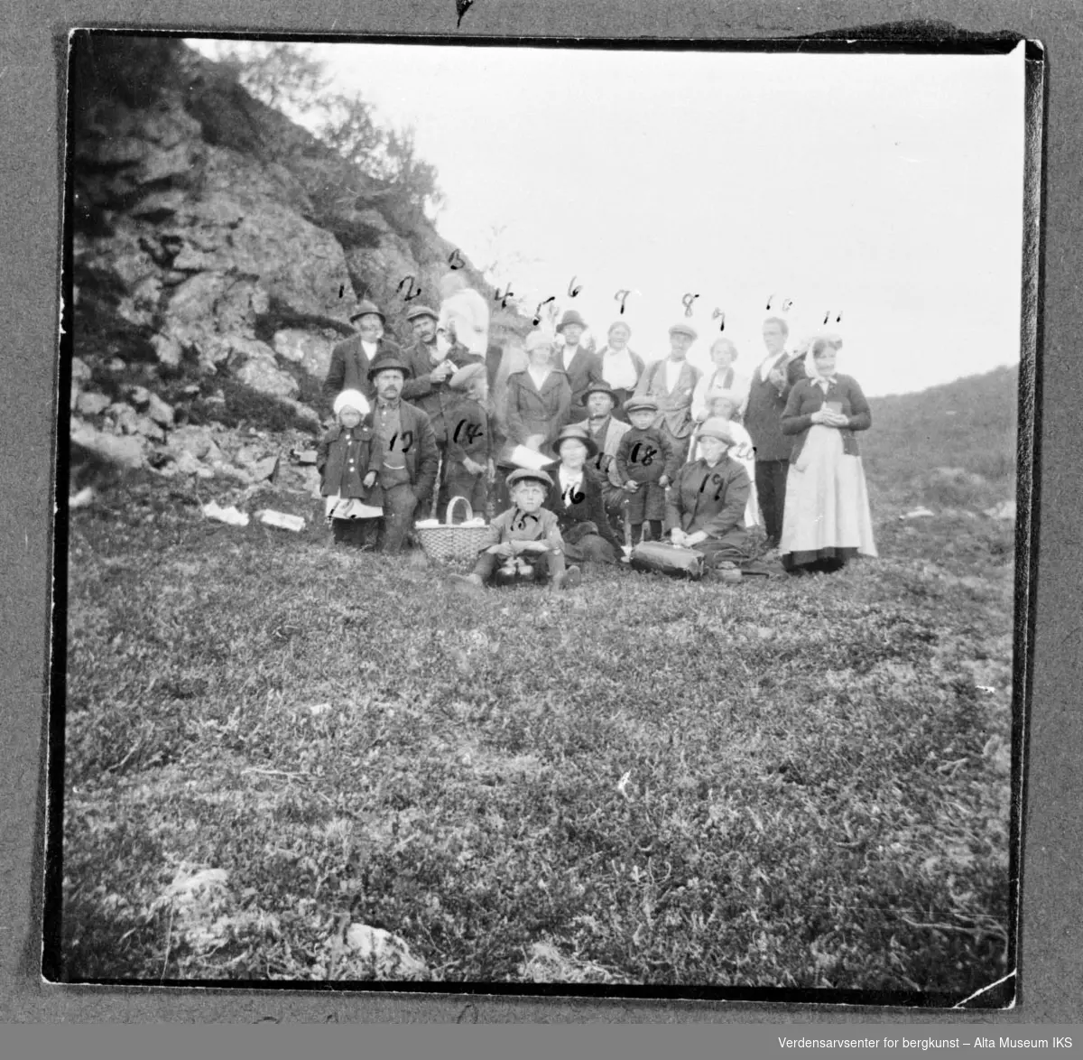 20 personer deltar på sankthansfeiring ved Kjerringvannet i Kviby 1922, på bildet ser vi flere voksne og barn med en nistekurv og fjell i bakgrunn. En mann til høyre ser ut til og holde en gitar.