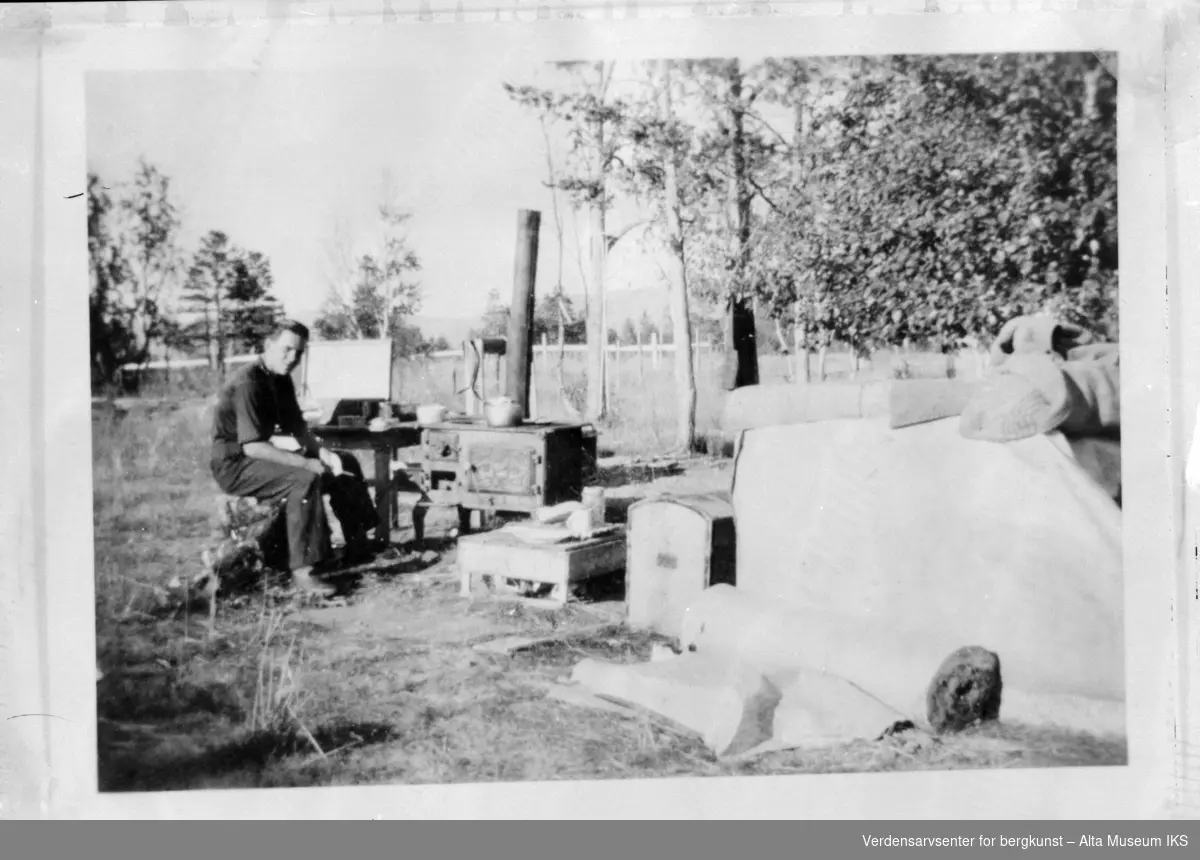 Bildet viser hjemkomsten til Saga etter krigen i 1945, på bildet ser vi en mann som sitter vedsiden av en ovn med flere eiendeler til høyre på bildet.