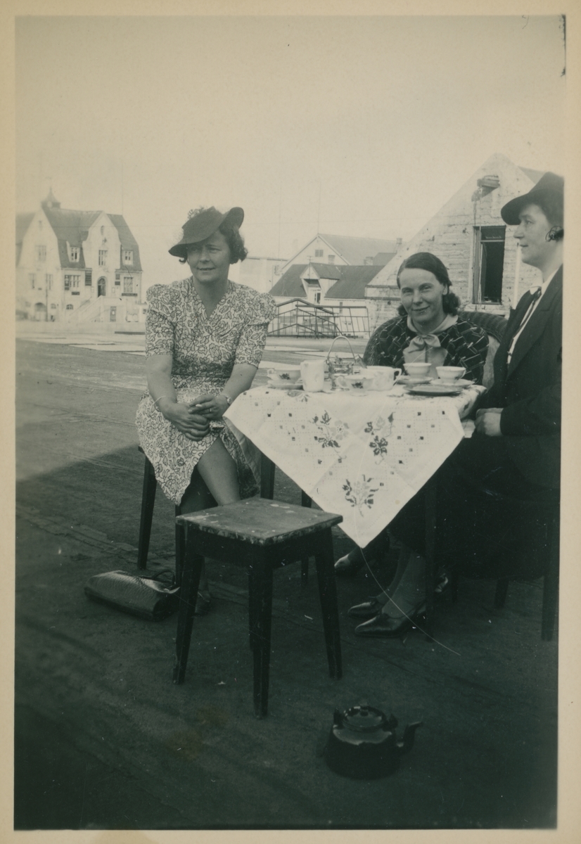 Tre kvinner drikker kaffe utendørs. Ekspedisjonsbygningen på dampskipskaia til venstre i bakgrunnen.