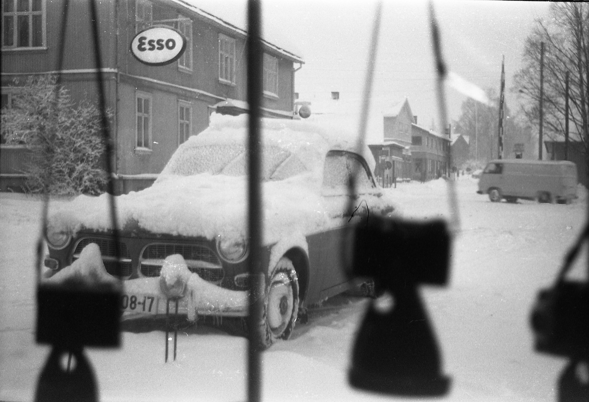 Snørikt gatebilde fra Lena, muligens vinteren 1962/63. Bildet er tatt gjennom vinduet i Lena Foto & Radio i "Raubua", i retning jernbaneovergbangen. Rett på utsida av vinduet står en delvis nedsnødd bil.
