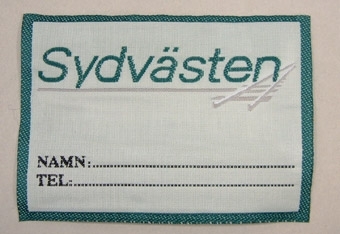 Tygmärke med invävt text i blågrönt på vit botten, samt grå räls (Sydvästens logotyp) och grön kant.
Under logotypen finns invävda rader för att skriva namn och telefonnummer.