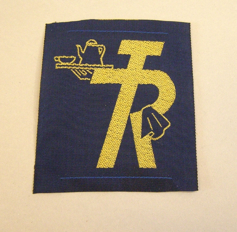 Rektangulärt tygmärke med mörkblå botten.
Maskinvävt logotyp med guldfärgad tråd: "TR-pojken", hållande en bricka med kanna och en kaffekopp.