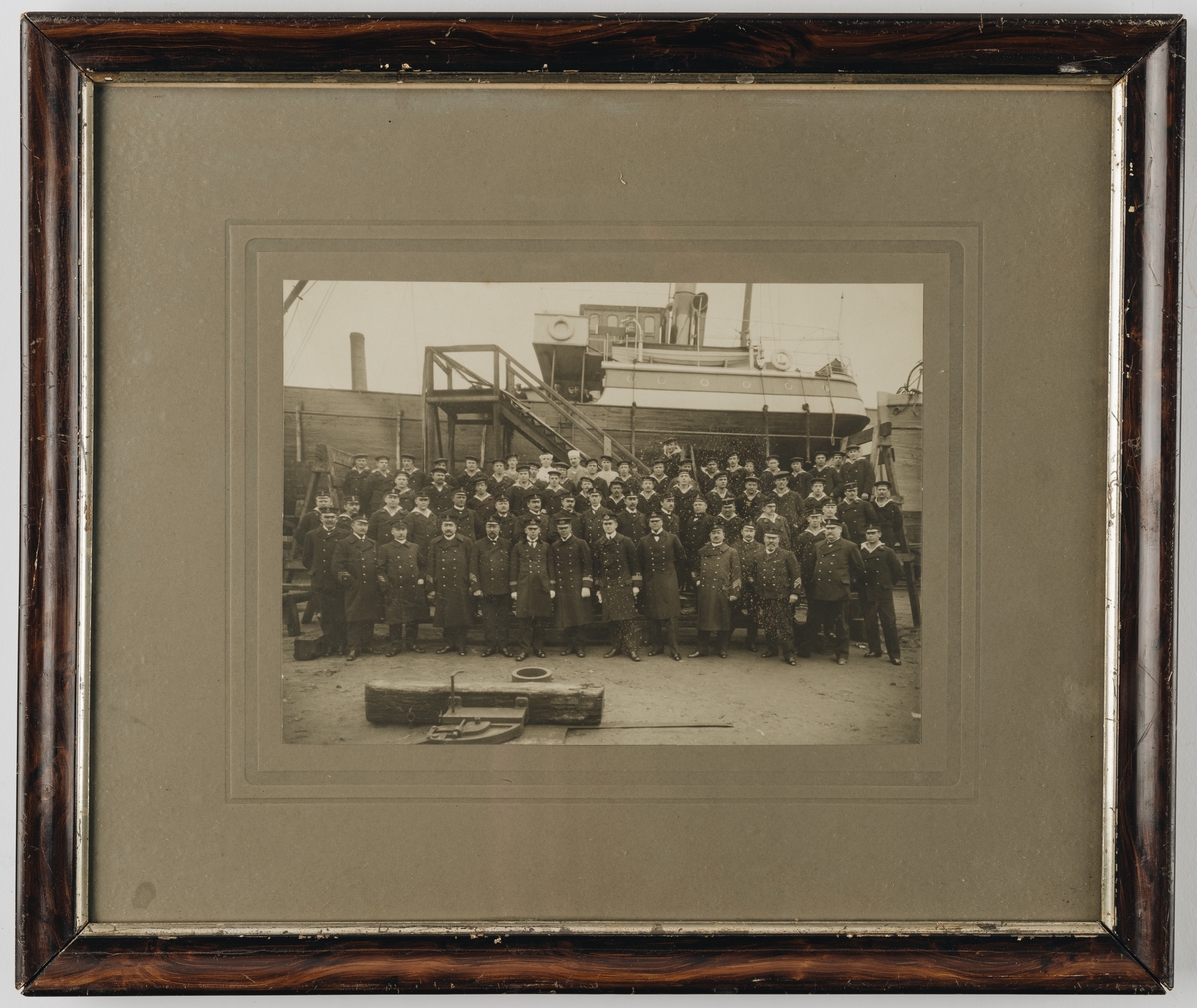 Bilden visar en grupp officerare, underofficerare och manskap från Svenska Flottan som har tagit uppställning för gruppfotografering framför ett träfartyg som ligger på land i ett hamnområde.