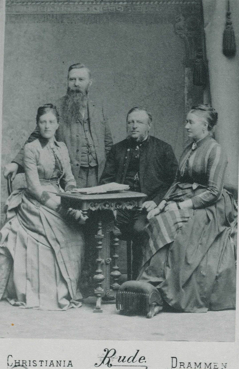 Familiegruppe med to kvinner og en mann som sitter ved et bord og en mann stående bak kvinnen til venstre i bildet.