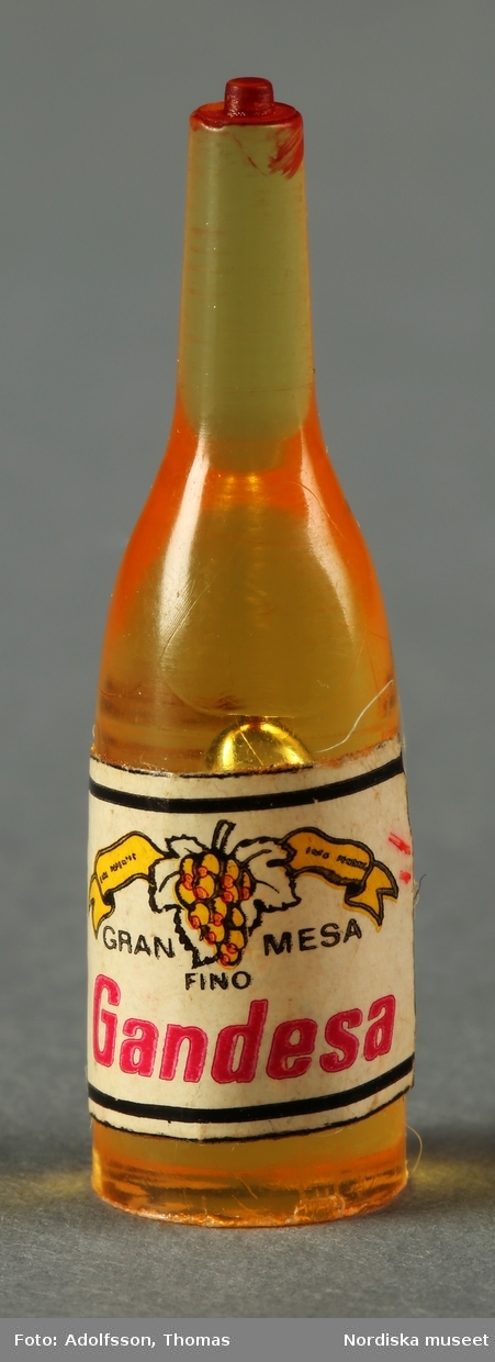 Tio flaskor som ska föreställa saft, drinkmix samt vin och sprit. Flaskorna är i olika färger som a-d) rött (4 st), e-h) gult (4 st) och i-j) grönt (2 st) och har små etiketter av papper. Hör till dockskåpsinredningen i matkällaren till dockskåp NM.0331721+.