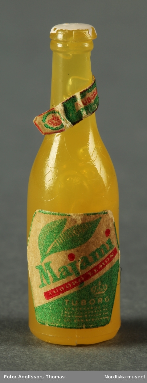 Tio flaskor som ska föreställa saft, drinkmix samt vin och sprit. Flaskorna är i olika färger som a-d) rött (4 st), e-h) gult (4 st) och i-j) grönt (2 st) och har små etiketter av papper. Hör till dockskåpsinredningen i matkällaren till dockskåp NM.0331721+.