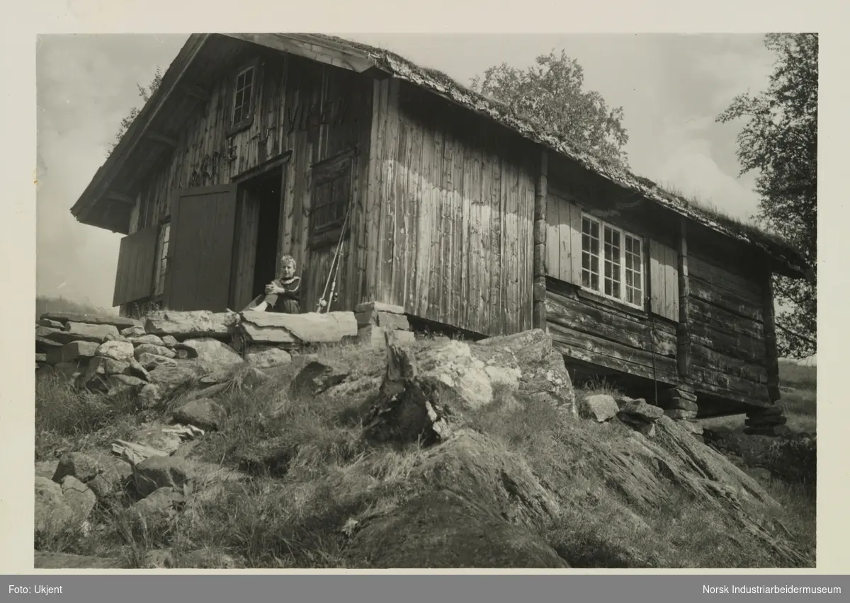 Person med to fiskestenger sitter utenfor hytte på Vermdeviki på Møsstrond.  Hytta er lafta med torvtak og over døren står på skriften "Varme-Viken"