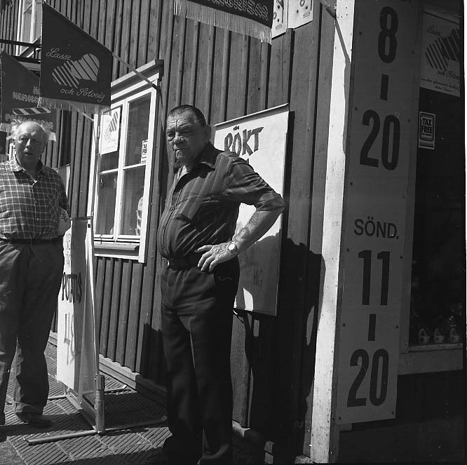 Två män står utanför en butik på Brahegatan. Det är Ingvar Åsbringer till vänster och Johan Arnell. Det är sommar.