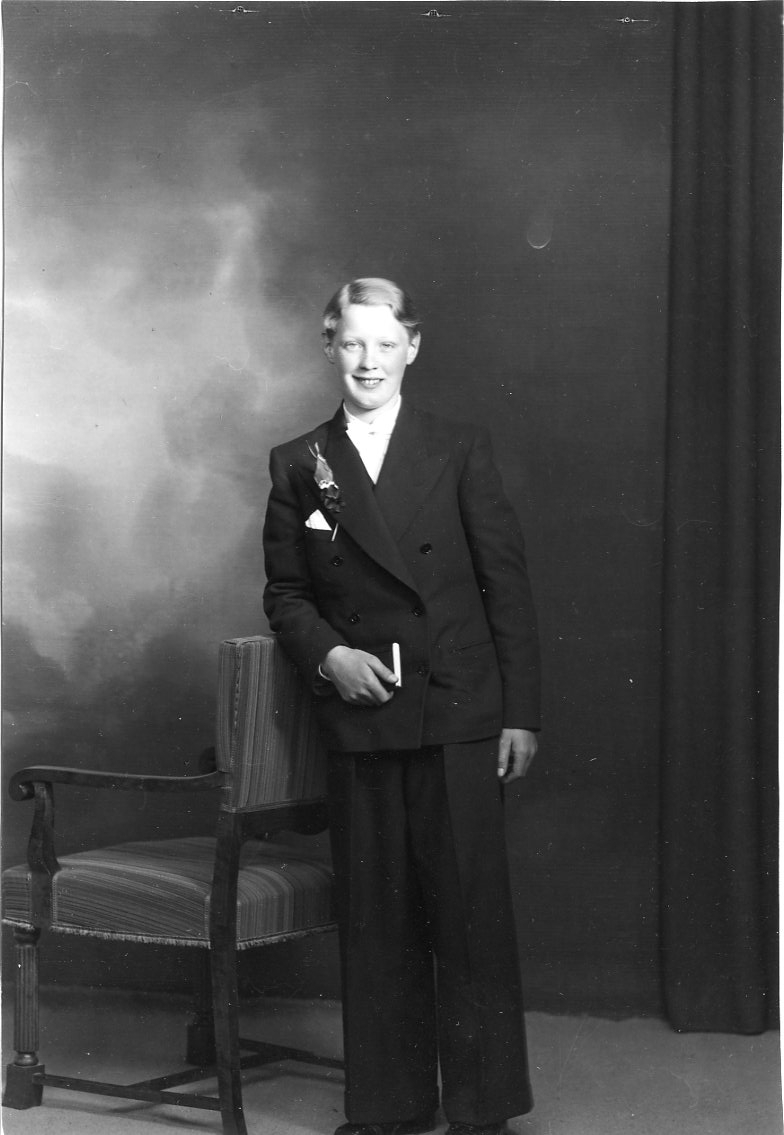 Porträtt av konfirmanden Kurt Johansson från Hövik. Han bär en mörk dubbelknäppt kostym, ljus skjorta med fluga samt en näsduk i bröstfickan. Möjligen en blomma på kavajslaget. I handen håller han en psalmbok.