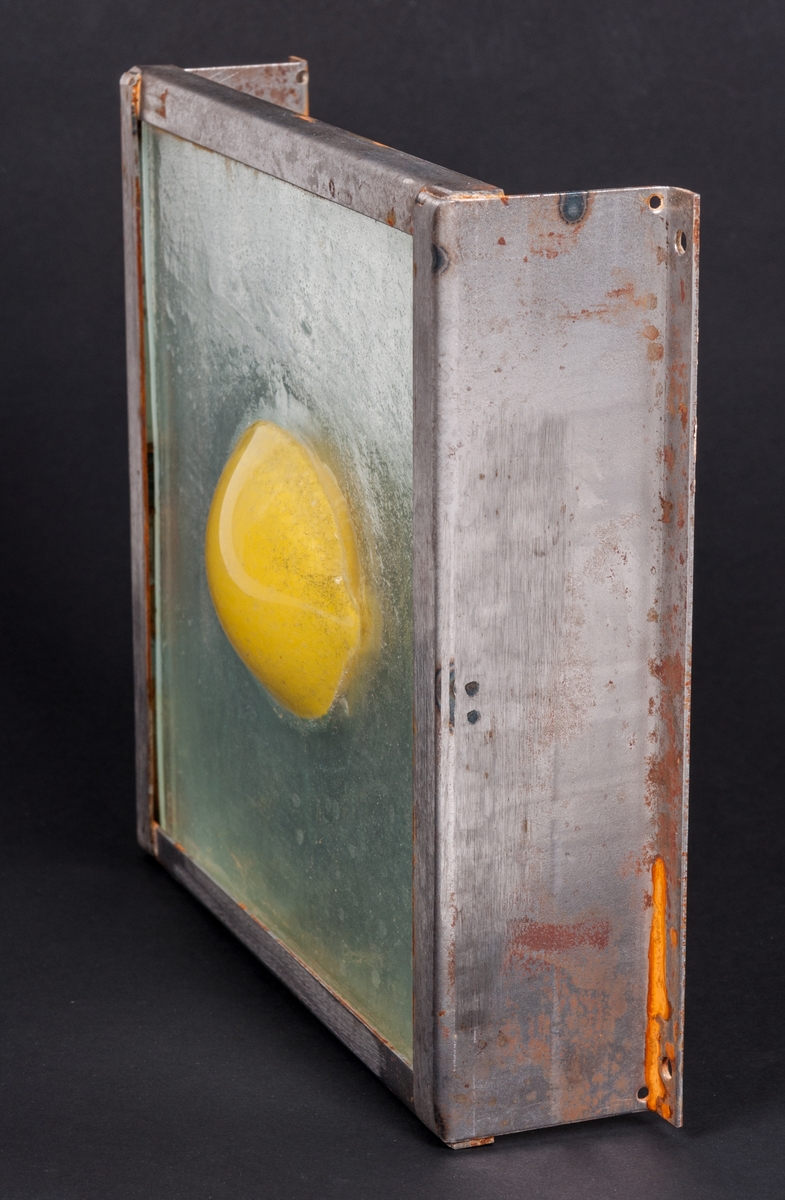 Objekt, skulptur, i glas och stål, "Spegelbild" (alt. Gul citron) av Lena Forslund-van Leer. Kvadratisk, något strukturerad, klar glasskiva med gul citron i relief i centrum. Bakom glasskivan är ett spegelglas. Glasskivorna infattade i obehandlad stålram.