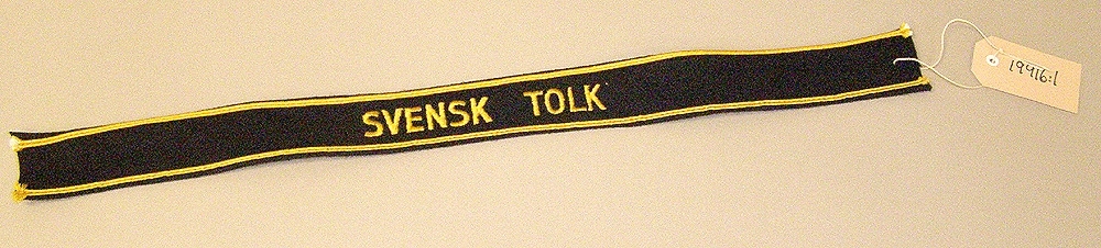 Mössband av svart bomull med rand uppe och nere samt texten "Svensk tolk" i gult.