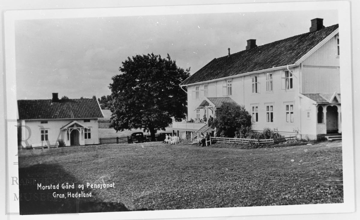 Tunet på Morstad vestre med en stor og en liten bygning. På verandaen og på bakken foran huset sitter det folk. En bil er parkert ved et stort tre.
