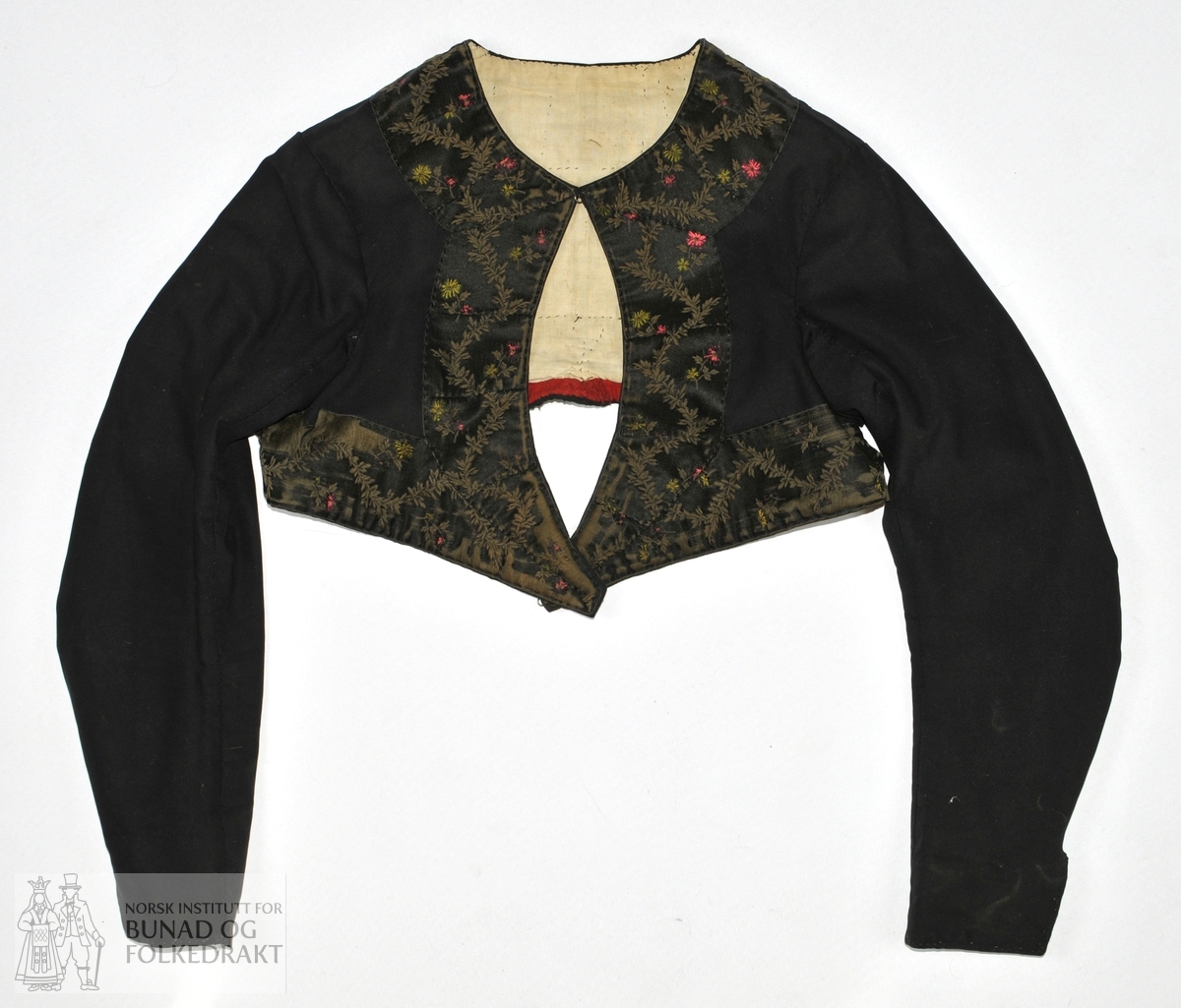 Kvinnetrøye i svart klede med fôr i ubleket bomull (toskaft). Trøya er kanta med et 7 cm bredt svart silkeband med mønster i rødt, grønt og brunt. Nederst på ermene 7 cm splitt. Sjå skisse.