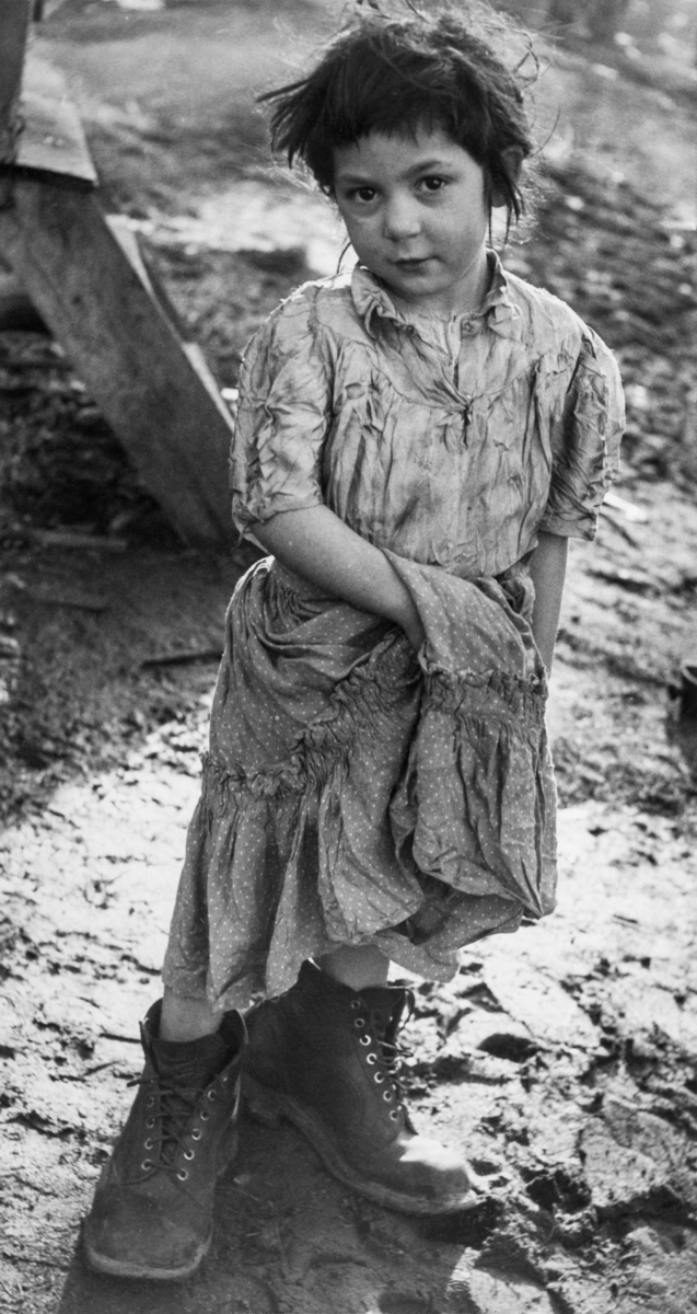 En flicka iförd klänning och för stora skor står framför ett tält på lerig mark. Troligtvis är bilden tagen i Lilla Sköndal.