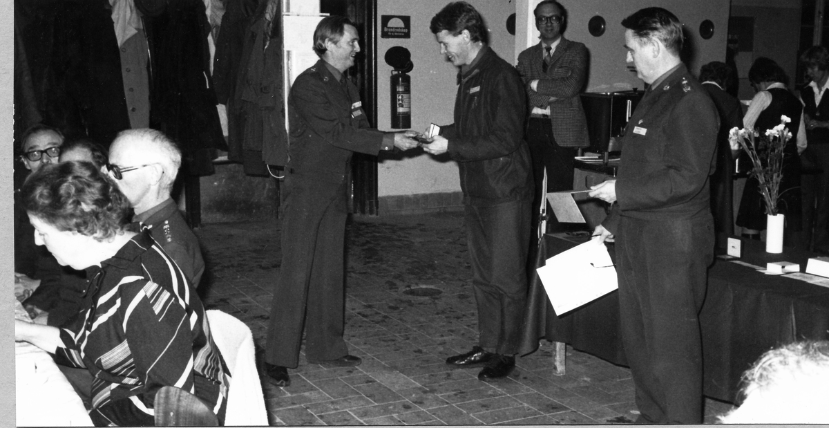 Samling med regementets personal 1979-12-18.

Öv 1. Åke Eriksson delar ut medalj till mj Sten Bråkenhjelm efter hans tjänstgöring vid I 19 och Pansartruppskolan (PS), övlt Nils-Göran Ahlgren assisterar.