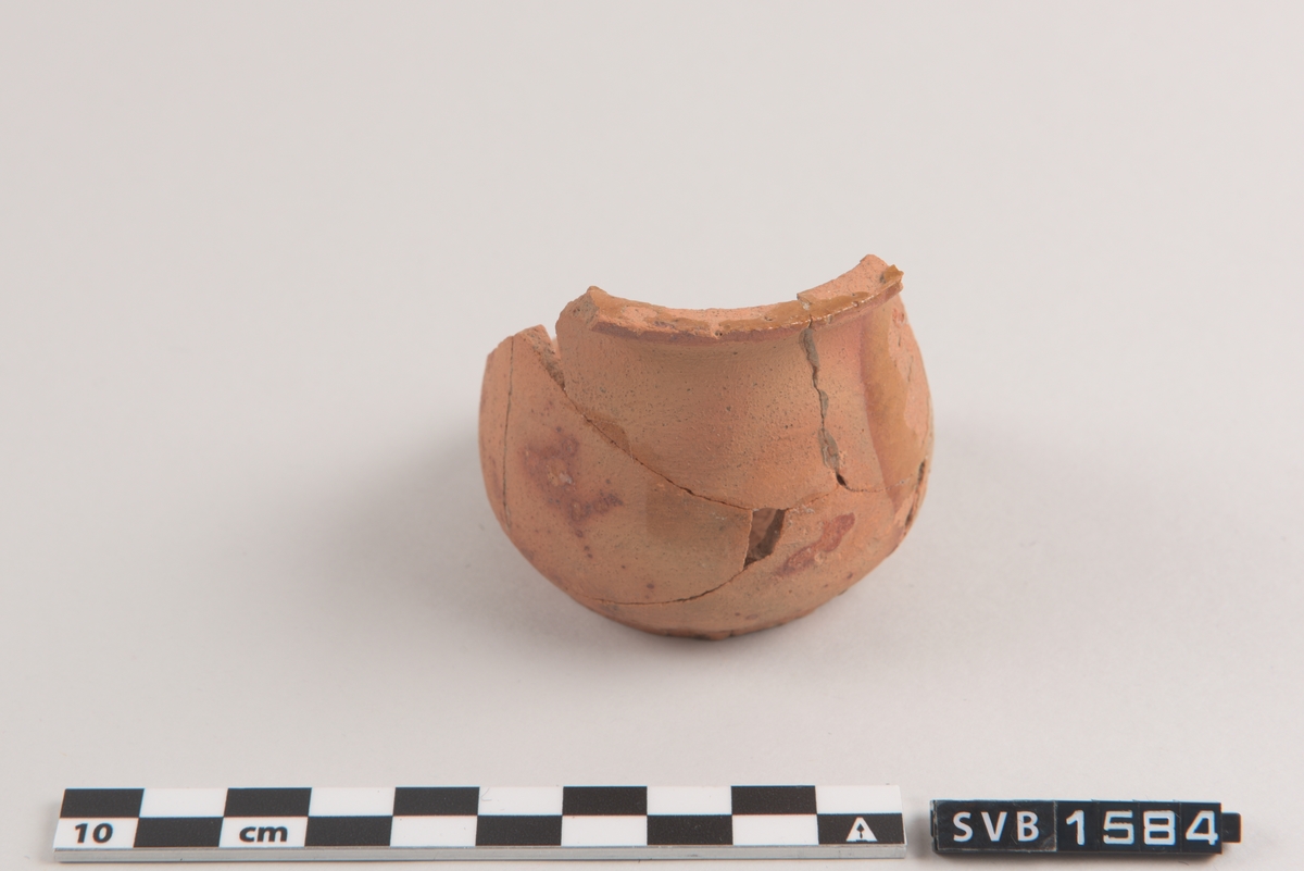 Terracotta-farget keramikk kopp. Bredere nedentil. En kant øverst. Koppen er ikke hel, men er limt sammen av fragmenter. Spor etter glasur.