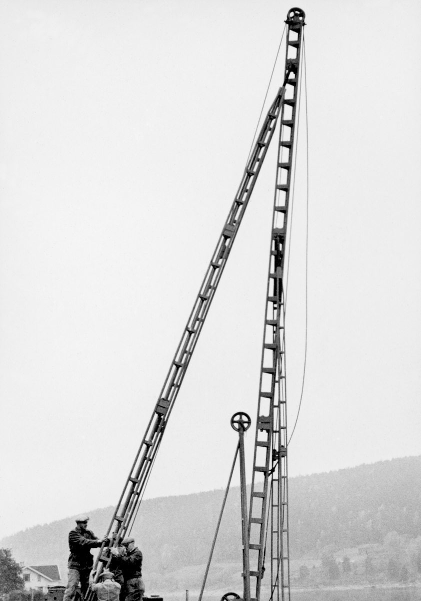 Reising av en rambukk på elvebrinken ved Furuholmen i Varteig høsten 1953.  Fotografiet er tatt mot ramma for den nevnte rambukken, som i dette tilfellet var tatt på land, der den skulle brukes til å drive ned en del pæler i ei fylling av leirholdig masse mot elva, for å forebygge utrasinger.  Bildet er tatt i forbindelse med reisinga av «kongen» – tårnet eller masta loddet på rambukken skulle heises opp i.  Tre karer later til å ha vært i ferd med å sikre det bakre støttebeinet på konstruksjonen.  Bak dem, nederst til venstre i bildeflata, skimter vi en av gavlveggene på våningshuset på gardsbruket Glenne.