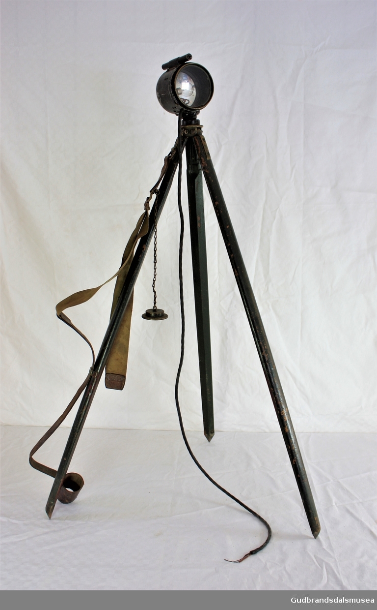 Signallampe med trefot stativ.Komplett med bærereim, med kabel som mangler stikker.
Brukt på Otta i Gudbrandsdalen under kampene der.
Lampa er merket 1918, og stativet London 1917.