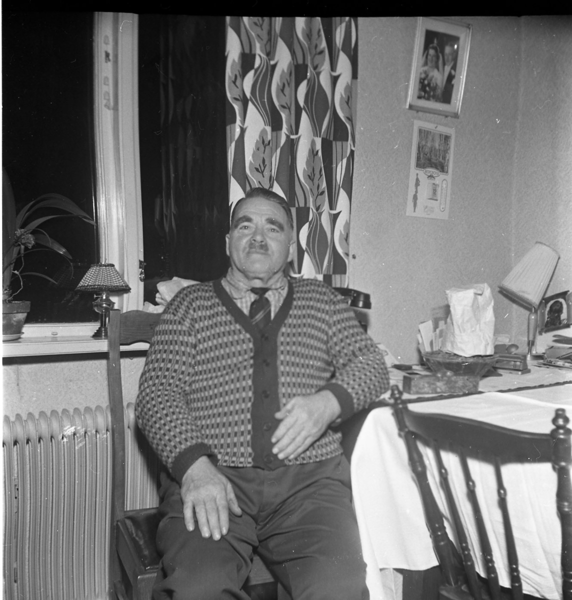 Porträtt av en äldre man som sitter på en stol vid ett bord med ljus duk, troligen på Sandviks gård. Han har mustasch, bär slips och en mönstrad stickad kofta. Han heter Gunnar Sääv.