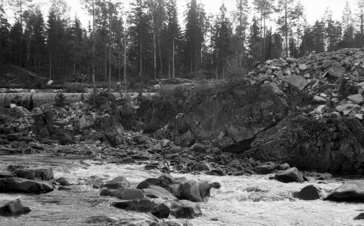 Fra elva Søndre Osa i Åmot kommune i Hedmark. Dette fotografiet ble tatt på tvers av elveløpet, like nedenfor Kvernfallet, der det i 1936 ble bygd en dam som skulle stuve opp vann til turbinen i en kraftstasjon som ble reist ved Brufallet, noen hundrede meter nedenfor. Mellom dammen og kraftstasjonen skulle det gå en kontinuerlig vannstrøm gjennom et såkalt turbinrør, som ved dette anlegget ble lagd av tre, omgitt av gjorder av jern. Vi ser en liten del av dette røret i den øvre delen av bildet. Ellers gikk deler av rørtraseen i ei grøft som var nedsprengt i berget. Det er masser etter slikt sprengingsarbeid som ligger på bergkollen til høyre i dette bildet.
