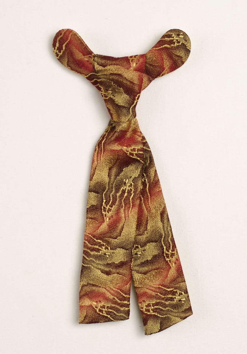 Kravatt av rött tyg med mönster i guld. Kort slips knuten och fäst på en halvcirkelformad hård pappskiva. Foder på skiva och slips av vitt bomullstyg.
Krok saknas.