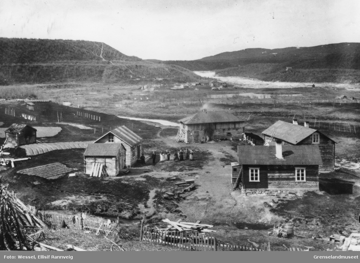 A. Wartiainens gård i Neiden i 1905. En gruppe mennesker står på gårdsplassen. I bakgrunnen ser vi skoltebyen med St. Georgs kapell på elvekanten og skoltenes boplass ved fossen.