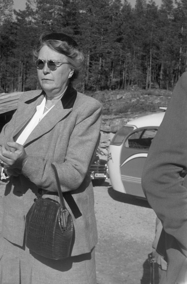 Ingeborg Magdalene Bødtker (1895-1985), fotografert under Norsk fløtningsforbunds befaring i 1952. Hun var gift med [Jacob Gottfrid] Otto Bødtker (1888-1966), som først var fløtingsdirektør i Drammensvassdraget (1915-1925), deretter i Skiensvassdraget (1925-1928). Følgelig hadde hun antakelig en slags vertinnefunksjon da organisasjonen ektefellen var direktør for samlet norske fløtingsfunksjonærer og styremedlemmene i Nordisk fløtningsråd til konferanse med innlagt befaring og årsmøte høsten 1952.  Ingeborg Bødtker var kledd i ei drakt med skjørt og jakke som på fotografiet framstår som lysegrå, med unntak av den bakre delen av kragen, som er mørk. Under jakka hadde hun en kvit bluse. Ingeborg Bødtker hadde alpelue på hodet, solbriller på nesa og håndveske over venstre underarm. Ellers var hun pyntet med perlekjede og perleøredobber. I bakgrunnen ser vi bakparten på ekskursjonsbussen og fronten på en bil av merket Plymouth, antakelig en årsmodell fra perioden 1946-1948.