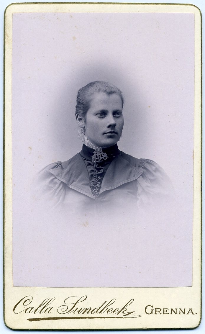 Kabinettsfotografi: porträtt av okänd ung kvinna med en brosch likt ett monogram vid halsen.