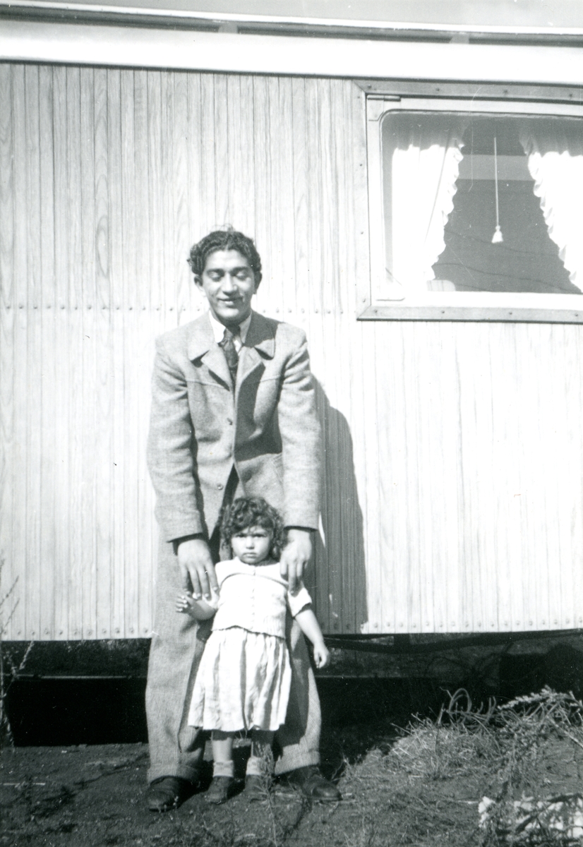 En ung man står tillsammans med en liten flicka framför en bostadsvagn. Bildens ursprung är okänt.