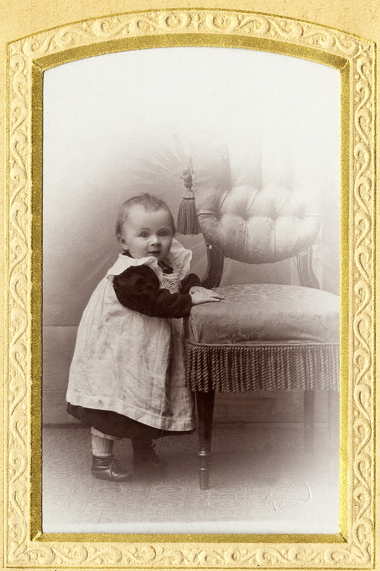 En liten flicka i kolt och förkläde som står vid en stoppad stol med frans. 
Helfigur. Ateljéfoto. 

Fotografens dotter.