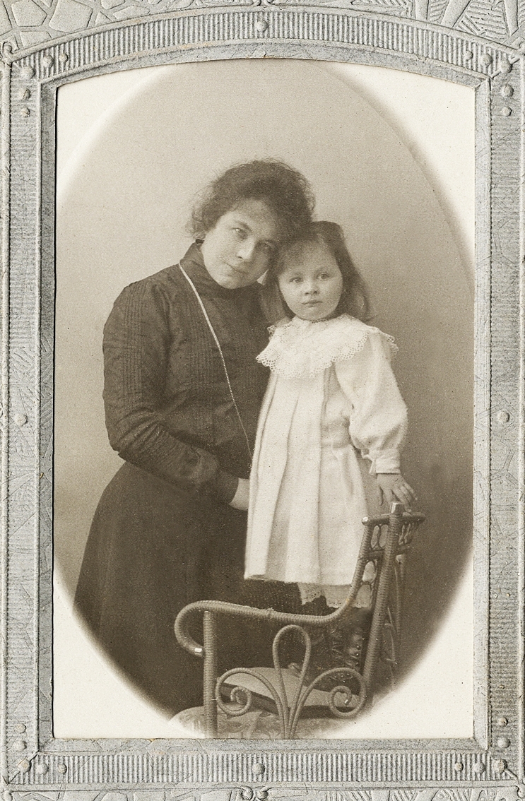 En kvinna i mörk klänning med hög krage och en liten flicka i ljus klänning, som står på en kudde i en stol. 
Knäbild. Ateljéfoto.

Fotografen med dotter.