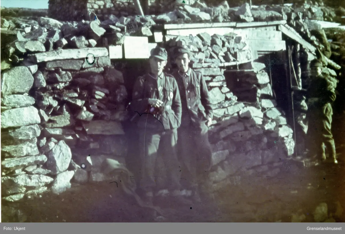 To soldater poserer foran en steinbygning. En tredje soldat står litt til høyre og åpner en dør.