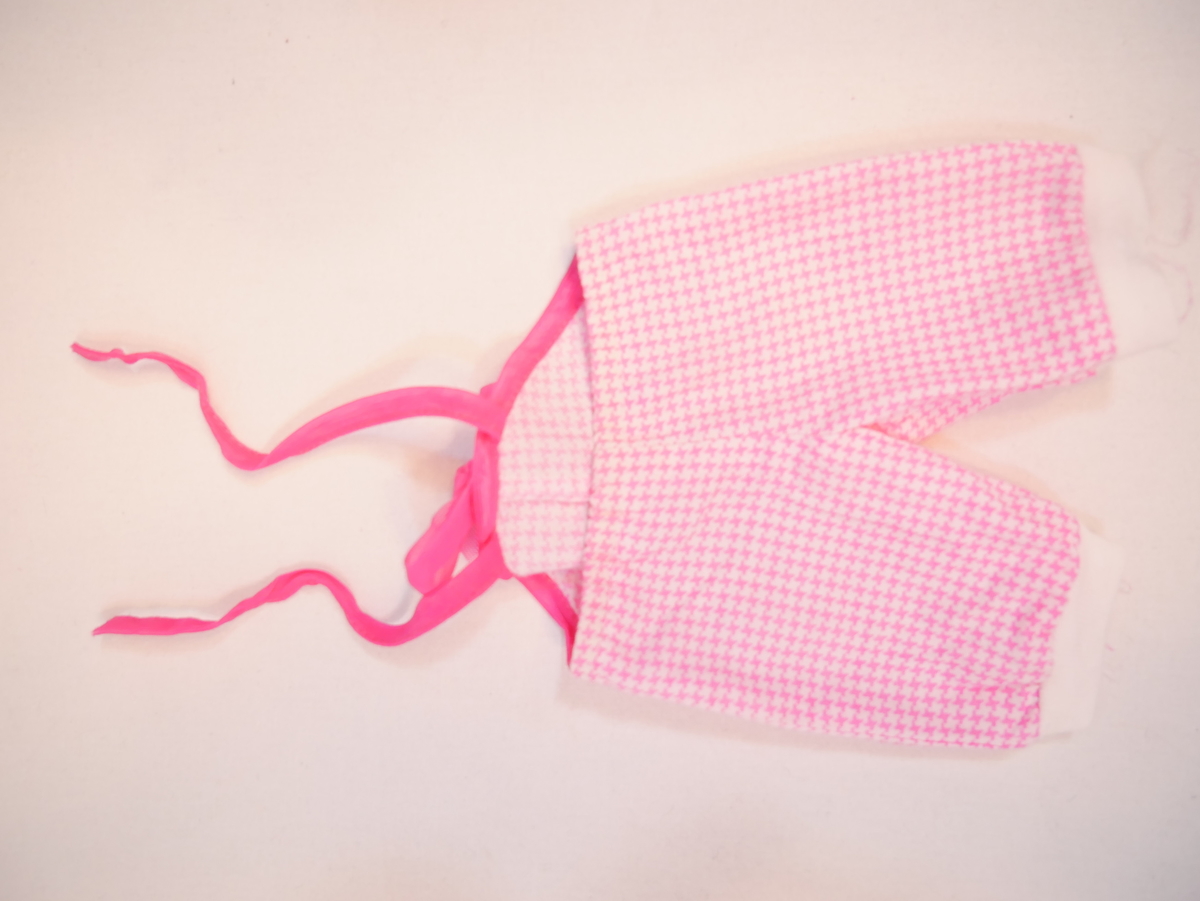 Dukkeklede som er kledd på SUM.09370. Dukkekleda består av ein kvit gensar og rosa sparkebukse. Gensaren har to trykknappar for opning/lukking bak. Ca 1/2 erme. Sparkebukse som går opp til brystkassa, med to rosa strikkband som knytast saman i nakken. Buksa er rosarutete, og har ei stor, rosa sløyfe på brystet. Kvite beinstrikk som er identiske med ermstrikka på gensaren.

Dukka vart gjeve til museet saman med fleire andre dukker med tilbehør og anna leikety (SUM.09360-09384).