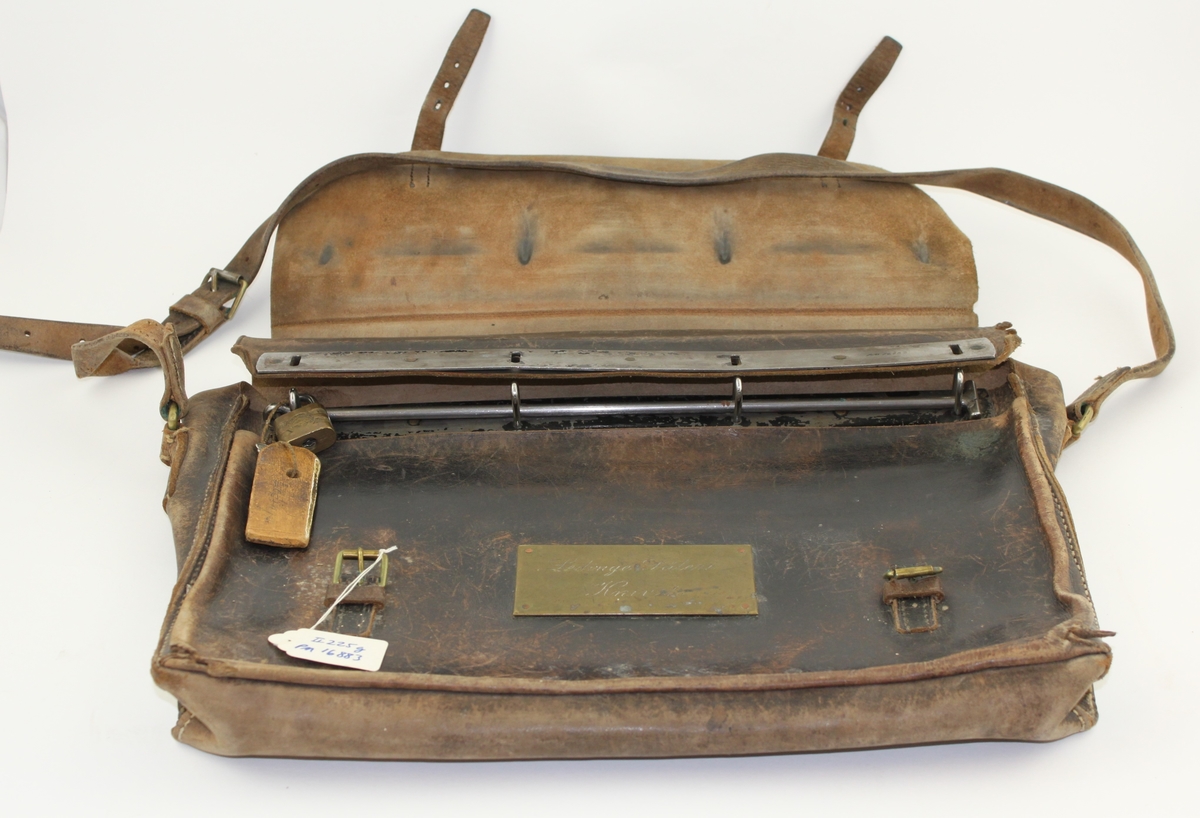 Lösväska i läder med klaff och två spännen. På framsidan en mässningsbricka med inskription i skrivstil. Väskan är försedd med låsten, hänglås och bärrem.