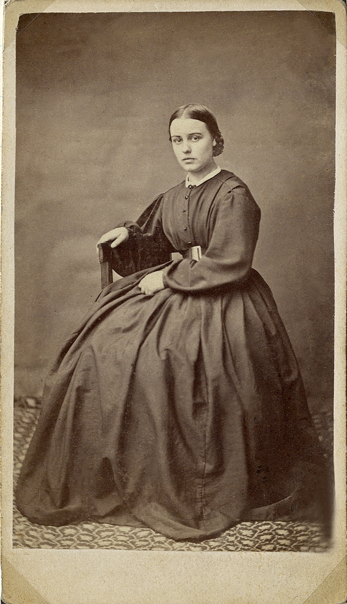 Porträttfoto av en ung kvinna i enkel mörk klänning med vit liten krage. Hon sitter på en stol och håller ena handen om ryggstödet.
Helfigur, halvprofil. Ateljéfoto.