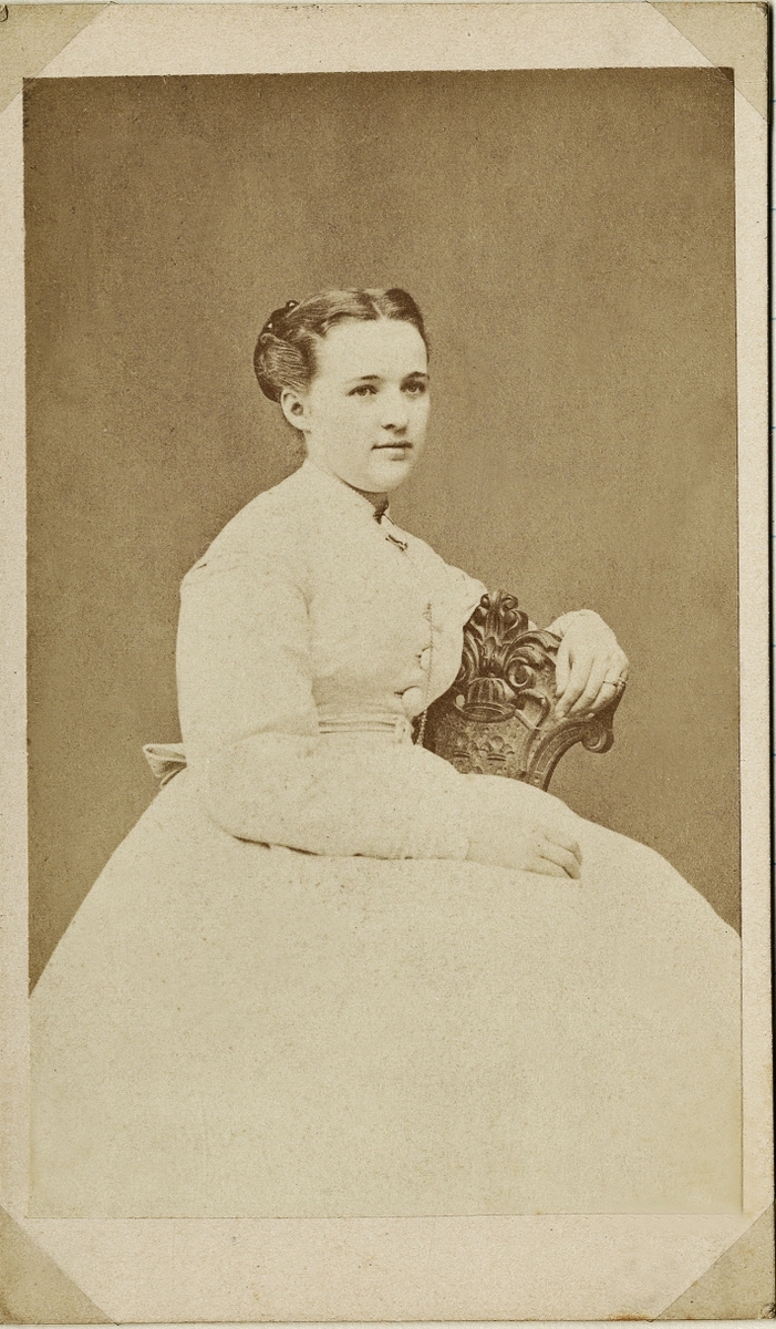 Porträttfoto av en ung kvinna i ljus krinolin, som sitter på en stol med snidat ryggstöd. 
Knäbild, halvprofil. Ateljéfoto.