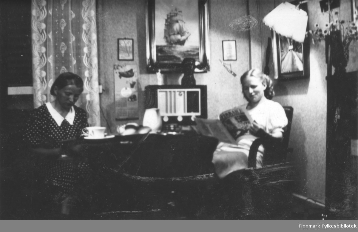 To damer sitter i stua og leser. Kaffekopper, boller i en fat og en kanne er på bordet. Radio i bakgrunn, noen tavler i veggen. Bildet er muligens fra Salttjern?