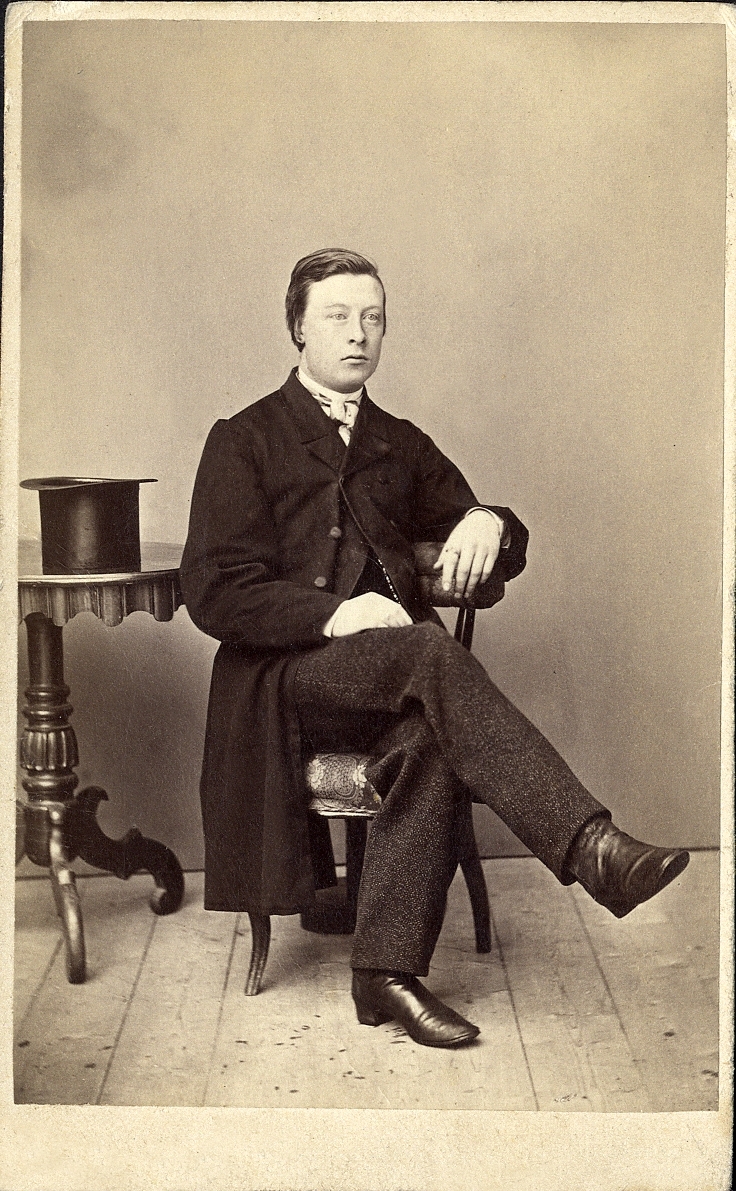 Porträttfoto av en ung man i bonjour med väst, stärkkrage och fluga. Han sitter på en stol vid ett pelarbord med en hatt på. 
Helfigur, halvprofil. Ateljéfoto.