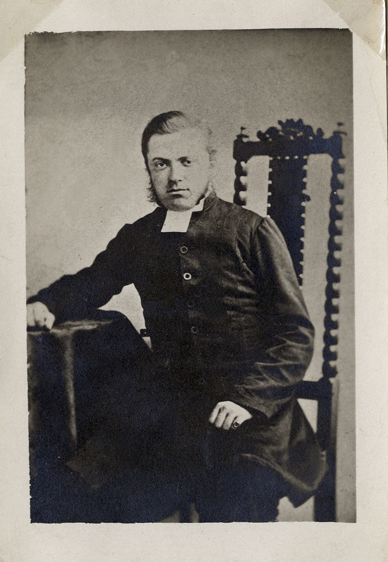 Porträttfoto av en man i prästrock med prästkrage. Han sitter på en högryggad stol vid ett bord med en mörk sammetsduk.
Knäbild, halvprofil. Ateljéfoto.