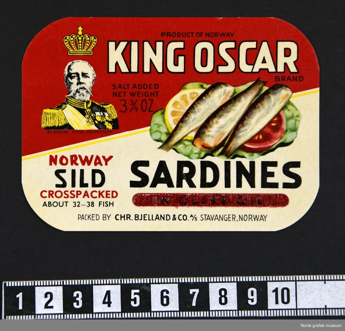 Rød og hvit etikett med en illustrasjon av sardiner dandert på salat, tomat og sitron. Logoen, portrett av Kong Oscar II, i venstre øvre hjørne. 

"Sardines in olive oil"