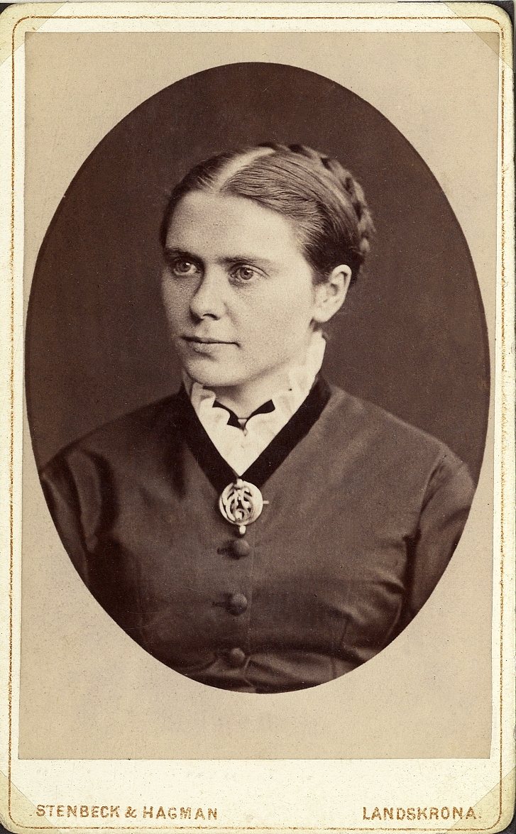 Foto av en kvinna i mörk klänning med sammetskrage och vit kråskrage. Vid kragen syns en brosch. 
Bröstbild, halvprofil. Ateljéfoto.