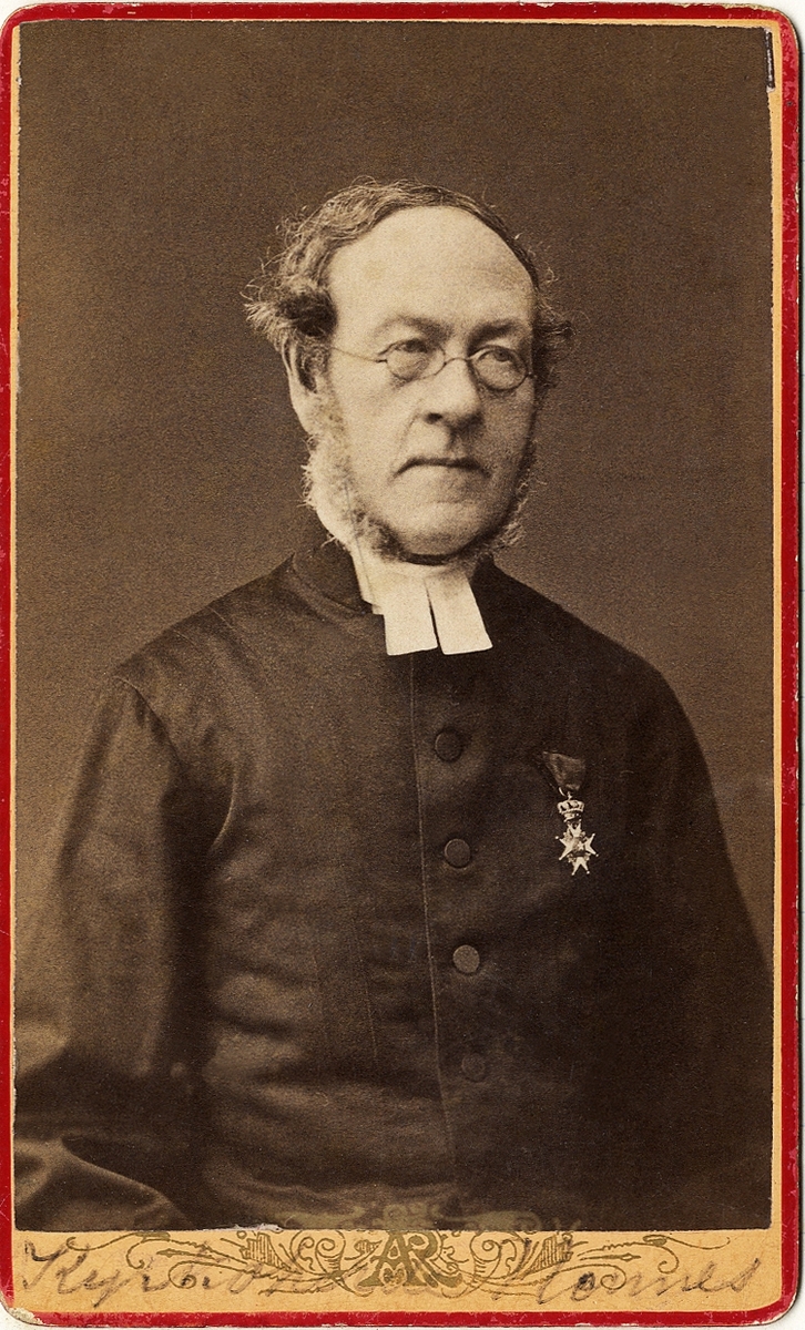 Foto av en man med glasögon, klädd i prästrock med stärkkrage och prästkrage. På bröstet syns Nordstjärneorden. 
Midjebild, halvprofil. Ateljéfoto.